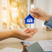 Persona entregando un llavero con forma de casa y el logo de Hogares ISN a un comprador, simbolizando la entrega de una vivienda. Sobre la mesa se observan documentos y un modelo de casa